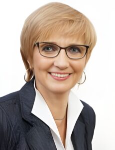 Frau mit blonden Haaren, Brille und schwarzem Blazer, lächelt in die Kamera.