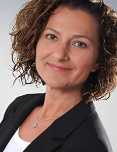 Porträt einer Frau mit lockigem Haar, die einen schwarzen Blazer trägt und in die Kamera lächelt.